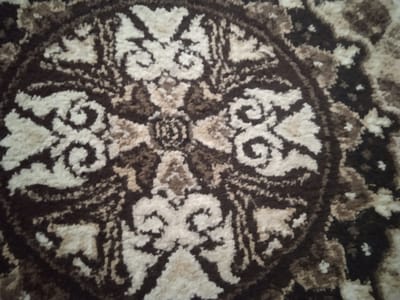 The image shows a close-up view of a carpet with a circular, ornate pattern. The pattern features swirls and floral motifs in shades of brown, beige, and white. The carpet appears to be made of a soft, textured material. There is no visible indication of the carpet's location within the city of Джапалак, Kyrgyzstan.