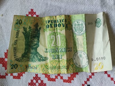 A Moldovan 20 lei banknote is laid out on a textured, white and red patterned cloth. The banknote features a portrait of Stephen III of Moldavia wearing a crown and armor. Text on the banknote reads "REPUBLICA MOLDOVA" and "DOUAZECI LEI". The denomination "20" is prominently displayed multiple times. The year "2015" and a serial number "D.0190" are also visible. The banknote is creased and appears to be made of paper. The cloth beneath it has a repeating geometric pattern in red on a white background.