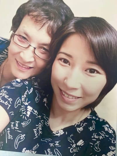 Two women are posing for a photo in an indoor setting. The woman in the foreground on the left is wearing glasses and a dark blue top with a white pattern of text and drawings. The text visible on her top includes "CHILL", "hey!", "hot", "stuff", "tuff", "hr", "hy!", "be", "pper", "baby", and "illy". She is smiling. The woman in the background on the right is leaning on the woman in the foreground and smiling. She has dark hair and is wearing a similar dark blue top with a white pattern. The lighting suggests the photo was taken during the day. The overall mood of the photo is warm and happy, suggesting a close relationship between the two women.