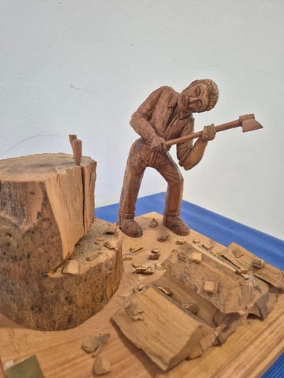 The image displays a detailed wooden carving depicting a wood-chopping scene, located in the city of El Espinar, Spain. The central subject is a carved male figure, rendered in light brown wood, wearing a shirt and trousers, with a beard and short hair. He is posed in a stooped posture, holding a simplified axe with both hands, positioned over his left shoulder as if mid-swing. To the left of the figure, a substantial, partially split wooden log or tree stump stands upright, with a section removed and two small wooden wedges inserted into its top surface. The carving's flat, rectangular wooden base is scattered with numerous small wooden chips, shavings, and several larger, irregularly shaped split logs, consistent with the activity of wood splitting. The entire carving rests on a blue surface, against a plain, light-colored wall.