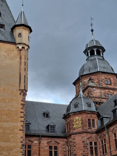 The image displays an architectural view of a historic building in Aschaffenburg, Germany, under an overcast sky. The scene is dominated by two distinct sections of a large structure.
On the left, a tall, slender, round tower or turret features a faded yellow-ochre plastered exterior. Its upper portion culminates in a dark grey conical slate roof, beneath which a small, rectangular window is visible. The right edge of this tower exhibits decorative vertical reddish-brown stone elements with carved detailing near its base, transitioning to a dark grey slate roof of an adjacent building section.
The right side of the image showcases a prominent section of a building constructed from reddish-brown sandstone blocks. A large, multi-tiered dome-shaped slate roof crowns a central tower, topped by a finial featuring ornate scrollwork and a small, pennant-like banner. Below the dome is an open belfry or observation level with arched openings. Further down, multiple rectangular windows with dark frames are set into the sandstone facade, which is adorned with intricate stone carvings and cornices. A golden, ornate shield or clock-like emblem, displaying radial lines and the inscription "1619" with an additional character, is mounted on a corner turret of this sandstone structure. Dark grey slate roofs with dormer windows are visible across various sections of the building. Greenish patina is present on some metallic architectural elements, suggesting copper.
The sky is a uniform, heavy grey, indicating overcast conditions. No people, animals, or active interactions are present in the frame.