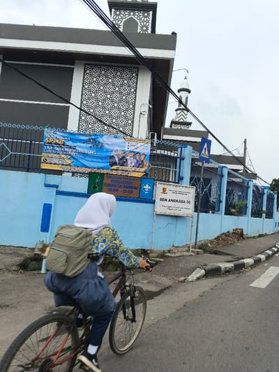 A young person wearing a white hijab, floral patterned shirt, and dark pants rides a bicycle on a street. The backdrop features a blue fence with a banner and a sign reading "SDN ANGKASA 06." A building with gray and white geometric patterns and a minaret rises behind the fence. Overhead power lines are visible against a cloudy sky.