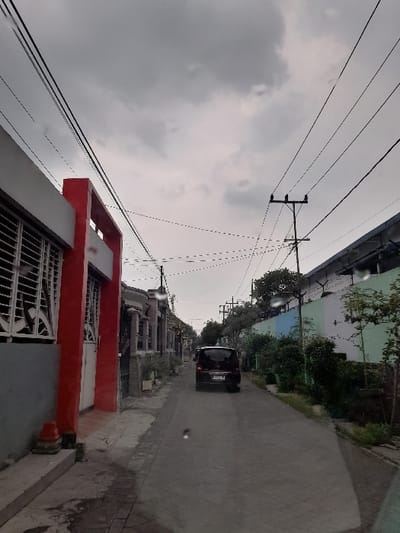 The image displays a street view in a residential area under an overcast sky, taken from what appears to be inside a vehicle looking forward. A paved road extends into the distance, with a dark-colored minivan or SUV driving away from the viewer in the middle of the lane.
Along the right side of the road, several houses are visible. In the immediate foreground, a house stands out with a striking red upper facade and a grey lower section, featuring decorative white grilles on its windows or gate. Further down the street, more residential structures are present, partially obscured by trees and bushes. The left side of the road also shows some greenery and building elements.
Above the street, a complex network of utility poles and electrical wires crisscrosses against the uniformly cloudy, grey sky. One prominent utility pole is visible towards the left, supporting multiple strands of wires. The weather appears to be overcast, with no direct sunlight, casting a subdued light over the scene.
No people are visible in the frame, and the street appears calm. The general architectural style of the houses and the overhead wiring are consistent with residential areas in Indonesia. No legible text is visible on vehicle license plates or any signs due to distance and image resolution. The presence of slight smudges or reflections, particularly along the edges, suggests the photo was taken through a window.