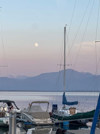 The image depicts a serene waterfront scene at dusk in Gstadt am Chiemsee, Germany. In the foreground, several boats are docked. Two sailboats, one with a blue sail and another with a green cover, are prominently featured. A motorboat with a tan cover is also visible, with two individuals standing on it. Another motorboat, partially obscured, is to the left, with two people near it.

In the background, a vast lake stretches towards a mountain range. The sky is a gradient of soft pinks and blues, illuminated by a full moon. The water reflects the dusky sky and the silhouettes of the boats. The overall atmosphere is peaceful and tranquil, suggesting the end of a day at the lake.