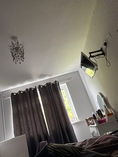 An interior scene, located in Fenland District, United Kingdom, depicts a bedroom viewed from a low angle, looking towards a window and an adjacent wall. The textured, off-white ceiling features a multi-tiered chandelier with crystal-like droplets in the upper left quadrant. Dark grey eyelet curtains, partially drawn on a metallic rod, frame a white-casement window through which bright green foliage is visible under daylight. A small white picture frame displaying "LOVE" text rests on the windowsill. The room's walls are light grey. A small, silver-framed flat-screen television is mounted on a black articulating arm on the upper right wall, connected to a white double power outlet. Below and to the right of the TV, a white dressing table with visible drawers stands against the wall, supporting an oval-framed mirror and several personal items, including a dark bottle, small containers, and sunglasses. The immediate foreground is occupied by light brown, wrinkled bedding, with a section of dark and light patterned fabric (possibly a sheet or pillow) visible at the very bottom. No people are present.