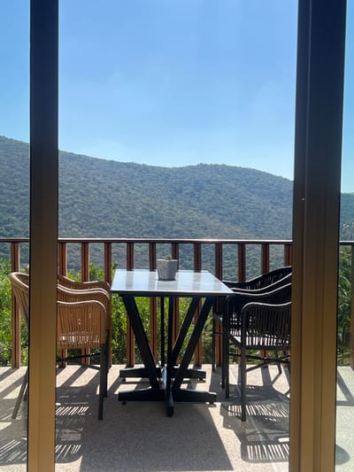 A patio view through a window features a table and chairs. The table, with a light-colored top and dark metal base, sits near a wooden railing. Two chairs are visible, one with a woven design and the other dark. Shadows from the furniture are cast on the patio. Beyond the railing, a mountain covered in lush green trees is visible under a clear blue sky. The window frame partially obscures the view on the left and right edges.