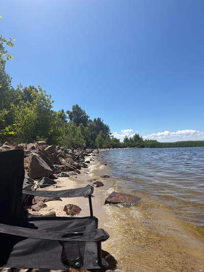 The image depicts a serene lakeside scene on a clear, sunny day in Lebedivka, Ukraine. In the foreground, two black folding chairs are positioned on a sandy and rocky shore, facing the water. The chairs are empty. The shore is lined with large, dark rocks, and further back, dense green trees and vegetation form a natural backdrop. The lake's surface is calm with gentle ripples, reflecting the bright blue sky dotted with a few fluffy white clouds. The water is shallow near the shore, revealing the sandy bottom and submerged rocks. In the distance, a line of reeds or tall grass marks the far edge of the lake. Two individuals are visible in the middle ground, standing near the water's edge. One appears to be an adult male, and the other, smaller figure, possibly a child, is partially submerged in the water. The overall impression is one of a peaceful, natural setting ideal for relaxation and outdoor enjoyment.
