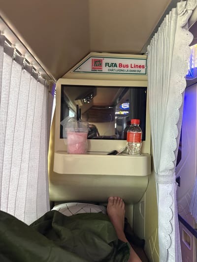 The image shows the interior of a sleeper bus bunk in Dĩ An, Vietnam. A person's bare foot is visible protruding from under a green blanket. On a shelf above, there is a clear plastic cup with a pink drink and two black straws, and a small bottle of water with a red label. Behind these items is a television screen reflecting lights. The bunk is enclosed by grey pleated curtains on the left and a white lacy curtain on the right. A logo for "FUTA Bus Lines" is visible at the top of the television area. The overall environment suggests a private sleeping compartment on a long-distance bus.