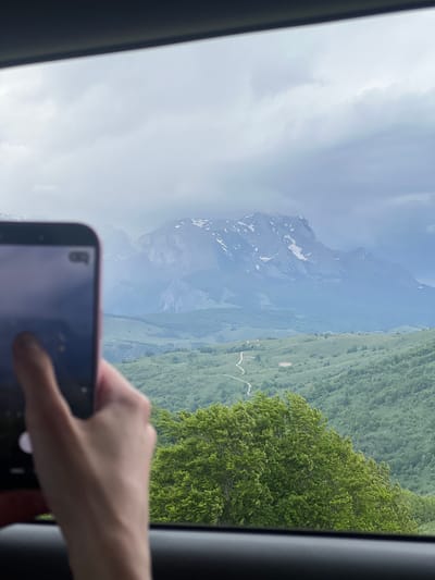 The image depicts a mountain landscape as viewed through a vehicle window. The foreground includes a hand holding a smartphone, partially obscuring the view. Through the window, a mountain range is visible with areas of snow cover. Adjacent to the mountains is a green, sloping hillside dotted with trees. It appears to be in the city of Pišče, Montenegro. The sky is overcast, indicating cloudy weather conditions. The presence of the window frame suggests the photograph was taken from inside a car or bus.