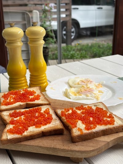 The image shows a still life of food items presented on a white wooden table, seemingly outdoors. The main focus is on the food, which includes two open-faced sandwiches topped with what appears to be red caviar and a layer of butter or cream cheese, served on a wooden cutting board. There is also a white plate with two poached eggs covered in sauce. In the background, there are two yellow wooden objects, possibly kitchen utensils.
Through a blurry reflection, part of a vehicle and some greenery can be seen, suggesting an outdoor or patio setting, with glimpses of the surrounding environment. The location is stated to be in the city of Tarasovka, Russia. There are no people visible in the frame. The overall composition suggests a carefully arranged breakfast or brunch setting.