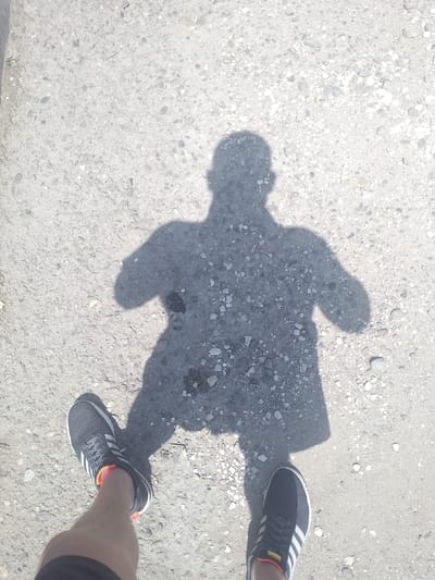 The image shows a shadow of a person standing on a rough, gravelly surface. The person is wearing athletic shoes, one of which is partially visible in the foreground. The shadow suggests the person is wearing clothing that covers their upper body. The sunlight is strong, casting a distinct shadow, indicating it is likely daytime. The scene appears to be an outdoor setting, possibly a pathway or road, given the ground's texture. The shadow's form and the strong sunlight suggest a sunny day, likely midday or afternoon. There are no visible text elements in the image. The location context of Ivanovskaya, Russia, provides a geographical setting for the scene.