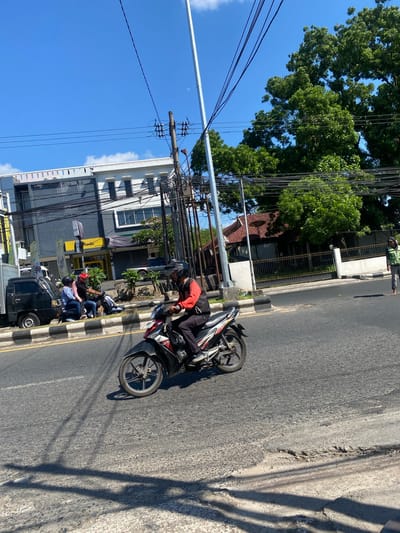 A sunny, bright day in Sungai Paring, Indonesia. The image captures a street scene with several elements. In the foreground, a motorcycle rider wearing a black helmet and a red and black jacket is seen riding on the right side of the road. The motorcycle is a dark color with red accents. Behind him, a black truck is parked on the left side of the road. Two people are seated on another motorcycle parked near the truck. The street has a white and yellow line marking and a raised curb.
In the background, there is a multi-story building with a gray facade and several windows. To the right of the building, a large green tree with dense foliage is visible. Power lines are strung across the sky, supported by utility poles. A small red-roofed house with a satellite dish is also visible further in the background, behind a fence. A lone pedestrian, wearing a green jacket and gray pants, is walking on the sidewalk on the right side of the road. The sky is a clear, bright blue with a few scattered white clouds. The overall impression is a typical day in a bustling Indonesian town.