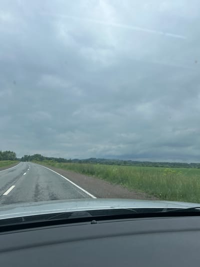 An overcast sky looms over a two-lane asphalt road that curves gently to the left, disappearing into the distance. The road is lined with a grassy shoulder and a strip of white markings. To the right of the road, a field of tall green grass stretches out, dotted with yellow wildflowers. In the background, a dense line of trees forms a dark green border, with rolling hills beyond. The foreground shows the dashboard and windshield of a vehicle, with the windshield wipers visible. The scene is devoid of people or any other discernible subjects. The image appears to have been taken from within a vehicle, likely in a rural or semi-rural setting in Kvarsa, Russia.