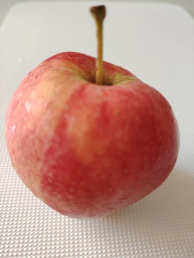 A single red apple with a short stem sits on a textured white surface. The apple is slightly off-center, positioned towards the bottom left of the frame. The lighting is bright, casting a subtle shadow to the right of the apple. The background is a plain, light-colored surface that appears to be a placemat or countertop with a raised, dimpled pattern. There are no people or other objects visible in the image, and no specific details suggest a location in Кривец, Russia.