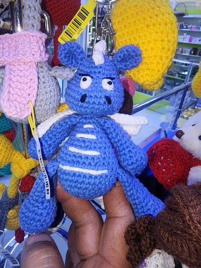 The image shows a close-up of a blue crocheted dragon keychain, held in a person's hand. The dragon has white wings and white accents on its body. Behind the dragon, other crocheted keychains are visible, including a yellow ball, a red item, and a pink and gray item. The background appears to be a retail environment, with shelves and products visible. The image is focused on the blue dragon and the hand holding it. No specific location in Ushtobe, Kazakhstan, can be identified from the image.
