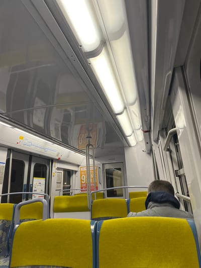 The interior of a subway car is visible. Bright fluorescent lights illuminate the gray, reflective walls. Yellow seats line one side, some occupied by a person with gray hair. Handrails are placed for standing passengers. A route map and signs are posted along the side, and doors are visible down the aisle. The image is taken at a tilted angle.
