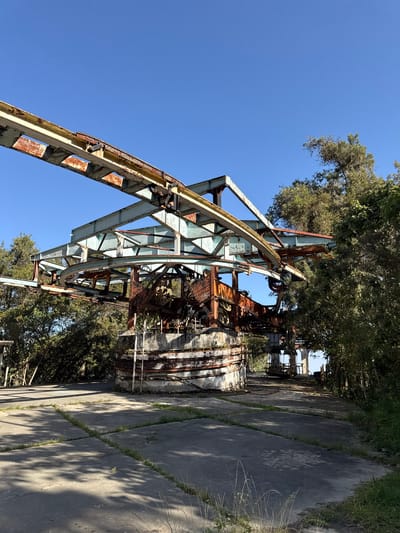 This image depicts the remnants of an abandoned, rusted metal structure in San Antonio de Galipán, Venezuela. The structure is complex, appearing to be an industrial-type construction with a skeletal framework of girders, supports, and circular elements. Rust is pervasive, indicating significant weathering and disuse. The structure is surrounded by trees and foliage, and it stands on a concrete foundation or surface with cracks and vegetation growing in the seams. The scene is bathed in sunlight, with a clear blue sky in the background. There are no people visible in the image. The focus is on the decay and abandonment of this once-functional structure.