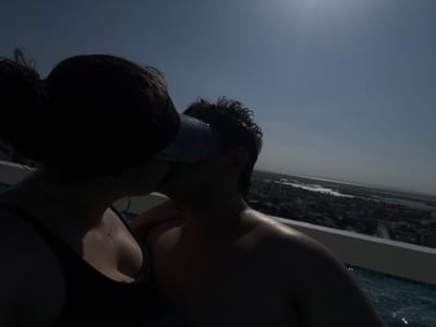 A low-angle, dark-toned photograph depicts two people, a male and a female, near a pool. The male is visible in the foreground, his hair and face facing the camera with a light-colored object near his lips. The female is positioned behind him, her dark hair partially obscuring her face. The photograph is underexposed, making it difficult to discern distinct features. Reflections of a city skyline are visible in the water of the pool. The location is possibly Tampico, Mexico, as indicated by a separate query.