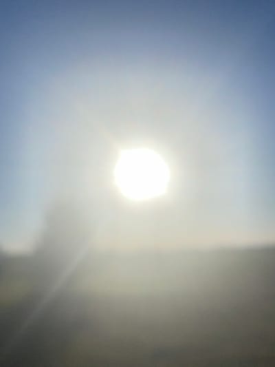 The image depicts a bright sun in a hazy sky, with a blurred landscape below. The sun is a luminous disc in the center of the frame, emitting rays that create a bright halo. The sky transitions from a pale blue at the top to a lighter, almost white hue towards the horizon, suggesting either sunrise or sunset, or a hazy atmosphere. The lower portion of the image is indistinct, showing blurred shapes and colors that suggest land and possibly vegetation, but no specific features are discernible. The overall impression is one of soft light and a muted color palette. The location is noted as Kozelshchyna, Ukraine, but the image itself does not contain any specific visual cues to confirm this location. There are no people or identifiable objects visible in the frame.