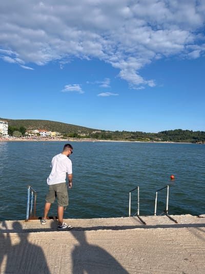 A man, viewed from behind, stands on a concrete pier overlooking the Aegean Sea in Potos, Greece. He is wearing a white t-shirt, olive-green cargo shorts, and athletic shoes. The man is looking down towards the water. To his left, a blue metal ladder extends into the sea. To his right, a metal ladder with four steps is partially submerged. An orange buoy floats in the water to the right of the ladder. In the background, a crowded beach with colorful umbrellas and buildings lines the shore. The sky is a clear blue with scattered white clouds. The scene is bathed in sunlight, casting shadows of the man and other figures onto the pier.