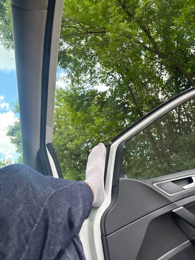 The image shows a person's legs and feet inside a car, with the car door open. The person is wearing grey socks and blue jeans. The viewpoint is from inside the car, looking up and out through the open doorway. Outside the car, lush green trees with dappled sunlight filtering through are visible, suggesting a daytime setting in a natural environment, possibly a park or a rural area. The sky above the trees appears to be blue with some white clouds, indicating fair weather. The interior of the car, specifically the door panel, is visible in the foreground, showing textured grey plastic and a chrome door handle. The overall impression is a relaxed moment, perhaps during a break from driving or while parked in a scenic spot. The location context indicates Pereiaslavske, Ukraine, but this specific detail is not visually evident in the image itself. No text is visible in the image.