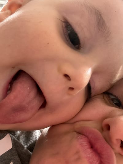 This is a close-up, low-angle, indoor photograph of two young children. The child in the foreground has their mouth open, sticking out their tongue. The child behind them has their eye visible, looking toward the camera. The image is tightly framed, focusing on the faces of the children. The background is dark and out of focus. The location of this image is the City of Yonkers, United States of America.