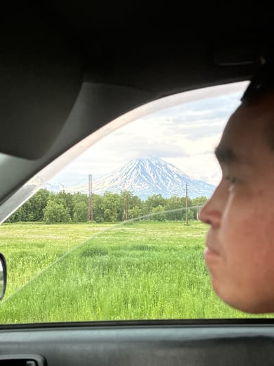 A young person's profile is visible in the foreground, looking out from the passenger side of a vehicle. Beyond the window, a vast, green field stretches towards a dense line of trees. In the distance, a snow-capped mountain dominates the horizon under a cloudy sky. Two tall, lattice-like metal structures stand within the field, suggesting the presence of infrastructure. The image appears to be taken from inside a car, with the dark interior framing the outdoor scenery. The specific location is in the vicinity of Razdol'nyy, Russia.