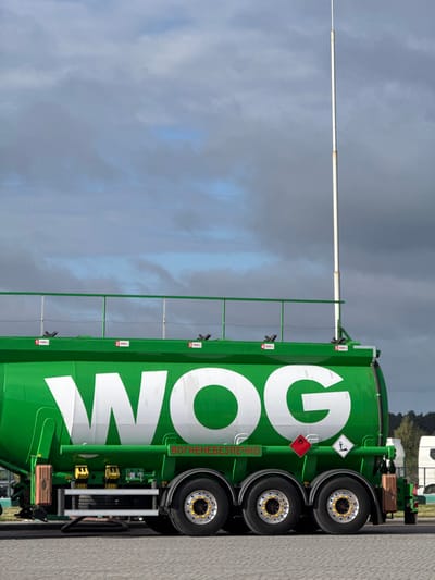 A bright green WOG fuel tanker semitrailer is parked on a light gray paved surface under a cloudy sky with visible patches of blue. The tanker prominently displays the white "WOG" logo on its side. Below the logo, a green banner with white text reads "ВОГНЕНЕБЕЗПЕЧНО" (FLAMMABLE). Further below, two diamond-shaped hazard placards are visible: a red one with a white flame symbol indicating flammability, and a black and white one with a tree and fish symbol indicating an environmental hazard. The tanker's top surface features a green metal access railing and five marked compartments, with capacities indicated as 11000 L, 8000 L, 7500 L, 5500 L, and a partially visible fifth compartment at 5500 L. A tall, slender white pole extends vertically from the forward section of the tanker, rising well above its structure into the sky. The semitrailer chassis visible features three axles with dual wheels, equipped with polished rims, black tires, and yellow lug nut indicators. Various mechanical components and a storage box are visible along the undercarriage. In the background to the right, a line of green trees and the white cabins of other commercial vehicles are partially visible. This scene is located at a commercial transport or fueling facility in Starovoitove, Ukraine.