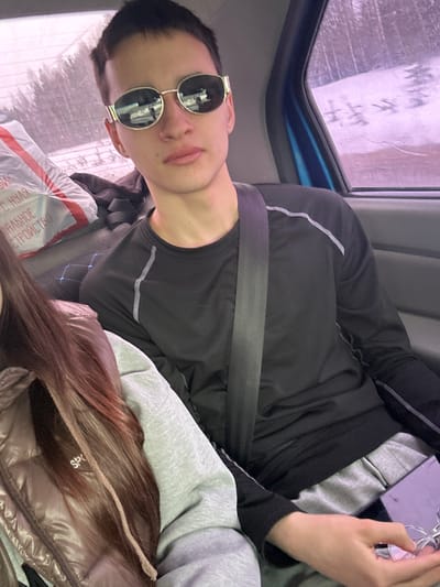 The image shows a person wearing sunglasses and a black shirt with a grey stripe, with a seatbelt across their chest, in what appears to be a vehicle. There's a window to the left. Another person's hair and part of a brown puffer jacket are also visible in the image. A package with Russian text is present. It is not possible to determine a location in the city of Ledukhi, Russia from the information given.