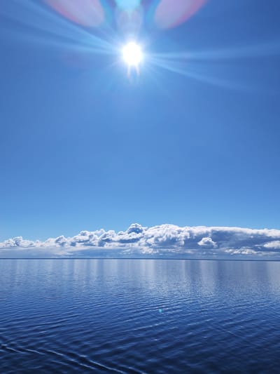 The image depicts a serene seascape under a bright, cloudless blue sky. The sun shines intensely, creating a lens flare effect at the top of the frame. A line of fluffy, white clouds sits near the horizon where the sky meets the water. The water is deep blue and slightly rippled, reflecting the light of the sun. The location is likely near Pavlokovo, Russia, based on the user's prompt.