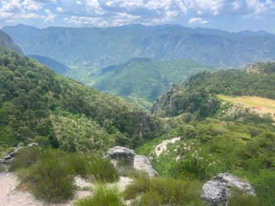 This is a landscape photograph taken from a high vantage point overlooking a valley and distant mountains. The foreground features a rocky area with sparse green bushes and grass. The middle ground is dominated by a steep, tree-covered slope leading down into a valley. The valley floor is dotted with vegetation and a winding path or road is visible. In the distance, a range of lush, green mountains stretches across the horizon, with layers of peaks fading into a hazy blue. The sky above is bright blue with scattered white clouds. The overall impression is one of a vast, natural, mountainous landscape on a clear day. The scene appears to be set in a rural or natural location, likely during daylight hours given the bright lighting and visible shadows. No people or specific objects are prominently featured, with the focus being on the expansive scenery. The location context provided is Klobuk, Bosnia and Herzegovina, which is consistent with the mountainous terrain depicted. There is no visible text in the image.