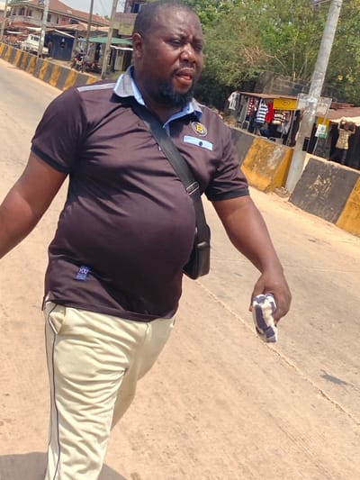 A male individual with a dark complexion, bearded, and of stout build, is captured mid-stride on a paved road in Afikpo, Nigeria. He wears a dark brown polo shirt featuring a light blue collar, placket, and a gold-colored "B" emblem on the left chest, with a small white tag reading "YOU" on the lower left torso. His lower attire consists of light beige trousers with a dark stripe along the side. A black messenger bag is slung across his left shoulder. In his right hand, he grips a folded, blue and white checkered cloth. His gaze is directed forward and slightly downward, with his mouth slightly ajar.
The environment indicates a bustling street scene. To the subject's right, a paved road surface shows cracks and seams, bordered by yellow and black striped concrete barriers. Beyond these barriers, several makeshift stalls or small shops are visible, displaying clothing and featuring corrugated metal roofs; one stall has a partially visible sign indicating "SANITARY STORE". A light grey utility pole stands upright near the barriers. In the deeper background on the right, additional indistinct figures are present, one appearing to carry an item on their head. To the subject's left, multi-story buildings, possibly residential or commercial, line the street, with a white vehicle partially visible near one structure. Lush green foliage and trees are present further in the background. The scene is illuminated by bright daylight, casting distinct shadows.
