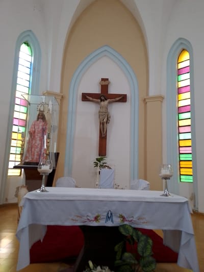 The interior of a church in Villa Hayes, Paraguay is shown. At the front is a white-clothed altar with two silver candle holders on top. Behind the altar, hanging on the wall, is a large crucifix. To the left of the crucifix is a glass case containing a statue of a saint or religious figure. On either side of the altar are arched windows with stained glass. The room has arched ceilings.
