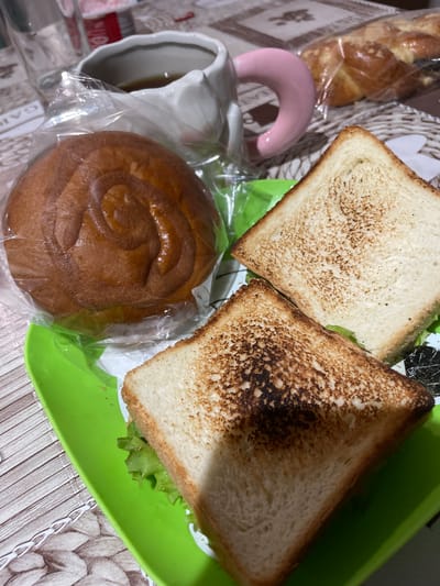 An overhead shot captures a breakfast spread on a table with a woven placemat. In the foreground, a bright green plate holds two triangles of toasted bread. The toast is golden brown with some areas slightly darker. Lettuce peeks out from between the bread slices of one sandwich. To the left of the plate, a round, swirled pastry is encased in plastic wrap. Behind the pastry and plate, a white mug with a pink handle holds a dark liquid, likely coffee or tea. In the background, partially visible, is a braided loaf of bread also in plastic. The table surface is visible beneath the placemat, displaying a patterned tablecloth. The scene is indoors, with no indication of the specific location within Zarichne, Ukraine.