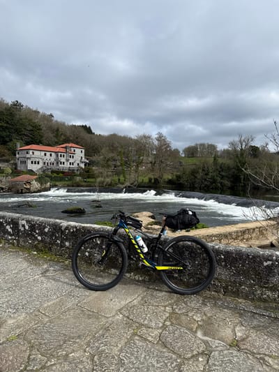 A Scott mountain bike, dark blue and black with prominent yellow accents, is positioned in the foreground, leaning against a weathered stone wall. The bike features disc brakes, a white water bottle in its frame cage, a small black top tube bag, and a larger black saddle bag. The stone wall, covered with patches of moss and lichen, forms the edge of a paved stone surface, likely part of a bridge or walkway.
Beyond the wall, a river flows with visible white rapids and small weirs stretching across its width, indicating a swift current. Sparse rocks and some minimal vegetation are present within the river. On the opposite bank, a large, multi-story stone building with a distinctive red tiled roof and numerous windows is situated on a hillside. This building is characteristic of the architecture found in the city of A Ponte Maceira, Spain. The surrounding landscape consists of hillsides covered with deciduous trees, many appearing bare, suggesting a dormant season. The sky above is overcast with a uniform layer of grey clouds. No individuals are present in the scene.