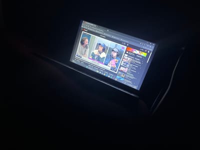 The image shows a tablet screen displaying a YouTube video titled "DESFILE de PERRITOS | QUÉ OLOR con Charo LÓPEZ, Noelia CUSTODIO y Mili GOGGIA GELATINA". The video features three individuals with headphones on, seemingly in a podcast-like setup. The YouTube interface with suggestions is visible on the left side of the screen. The tablet screen also displays a weather update from Llavallol, Argentina. The tablet is indoors, and the scene is dimly lit.