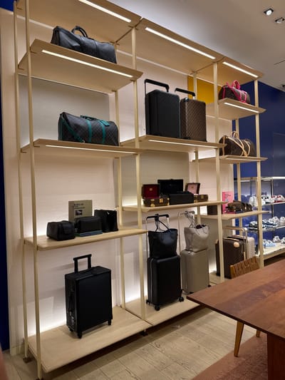 The image shows a display of luggage and bags inside a Louis Vuitton store. The merchandise is arranged on shelves within a light wood-colored display unit against a background with a blue section. The items include suitcases, duffel bags, and smaller bags. There is a wooden table edge visible at the top right corner of the image. The scene appears to be in a retail setting. The geographical location can only be inferred from the user's prompt, suggesting the store may be in Oak Brook, Illinois, USA.