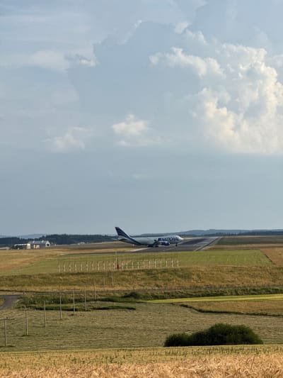 In Hahn, Germany, on a cloudy day, a National Airlines cargo plane is on the tarmac, its engines possibly engaged, suggesting it is either preparing for takeoff or has just landed. The aircraft, a large jet with "NATIONAL" printed on its side, is positioned on a runway that cuts through a landscape of green and gold fields. The surrounding area is characterized by rolling hills dotted with trees and some low-lying buildings, possibly hangars or airport facilities, in the distance. The foreground features fields of dry wheat, adding a rustic touch to the otherwise industrial scene of the airport. The sky is partially obscured by a mix of white and gray clouds, with patches of blue peeking through, indicating variable weather conditions.