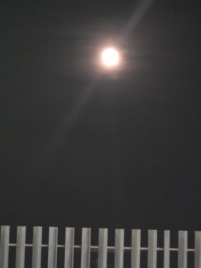 A low-angle shot features a bright, diffuse light source in the upper-left quadrant against a dark background, possibly depicting the moon or a strong artificial light. To the right, a series of horizontal slats, likely window blinds, extend vertically along the right side of the frame. The scene's darkness suggests an indoor or nighttime setting. The location in the city of Sant Andreu de Llavaneres, Spain cannot be confirmed from the visual information alone.