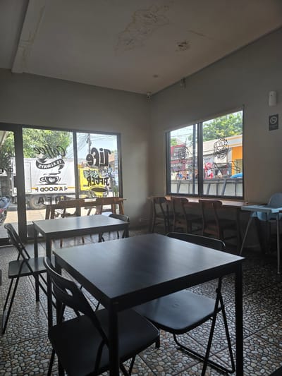 The image depicts the interior of a coffee shop with large windows looking out onto a street in Klapanunggal, Indonesia. The interior features several dark-colored tables and chairs, with a patterned tile floor. The windows have text and graphics applied to them, including phrases like "Coffee" and "Life is Always Better". Outside, a yellow truck and various buildings are visible. A "No Smoking Area" sign is posted on the wall. The ceiling shows signs of water damage and staining. There are no people clearly visible within the coffee shop in the foreground, but people can be seen outside the window. A child's high chair is present in the background.