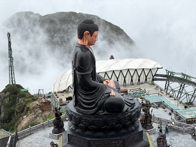 A large, dark-robed Buddha statue sits in a meditative pose atop a tiered lotus pedestal. The statue is the central subject, with its head and shoulders visible in profile. Behind the statue, a misty mountain range is shrouded in fog, with a cell tower visible to the left. In the middle ground, a large white structure with a geometric pattern and a green metal framework is partially obscured by clouds. A walkway with glass railings and several small buildings are visible below. The foreground shows a paved area with several smaller statues and people walking around. The weather appears overcast and foggy, suggesting it is likely morning or a cloudy day. The scene is set on a mountaintop, possibly a tourist attraction, given the infrastructure and presence of visitors. The overall mood is serene, amplified by the Buddha statue and the misty, atmospheric conditions. No visible text is present in the image.