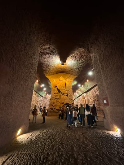 A horizontal, dimly lit, indoor shot depicts a statue or carving in Shizhong, China. The view is directed into a tunnel-like space with rough-textured walls. On the right side of the image, a row of individuals is standing facing the statue. Small lights are spaced along the tunnel's ceiling, casting a warm glow on the statue and surrounding walls. The statue is the central figure, detailed with flowing lines, and it appears to be part of a wall carving. The composition includes the statue, the people, and the interior architecture of the tunnel.