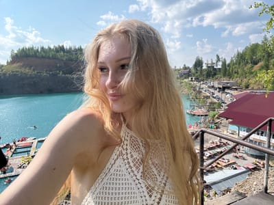 The image captures a woman with blonde hair wearing a white crocheted top, taking a selfie with a body of water and a recreational area in the background. The location appears to be Karyer Izvestnyak, Russia, a former limestone quarry transformed into a tourist destination.
In the foreground, the woman is positioned to the right, her face turned to the left, with her blonde hair flowing around her shoulders. She is wearing a white, intricately crocheted halter top. Her left arm is extended, presumably holding the camera.
The middle ground reveals a vibrant blue body of water, likely the quarry lake. Several wooden platforms and docks extend into the water, with people visible sunbathing and engaging in water activities. Some people are on paddleboards, and others are near boats.
Further in the background, the scene transitions to a bustling recreational area. There are numerous sun loungers and umbrellas spread across a sandy or gravelly area. Buildings with red roofs are visible, suggesting amenities for visitors, such as cafes or changing rooms. A parking area with several cars is also present, indicating visitor access. The environment is characterized by lush green trees and vegetation covering the slopes of the quarry, under a bright blue sky with scattered clouds. The overall atmosphere is one of a sunny, recreational day out.