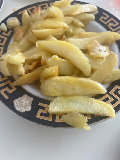 A close-up shot shows a plate of golden brown french fries with a decorative black and gold rim. The fries are piled high on the plate, with some slightly darker, crisped edges. The plate itself has a repeating Greek key pattern and a circular motif of a face, likely a Medusa head, on its rim. The lighting is bright, suggesting an indoor setting. The location is not identifiable from the image itself, and no information about the city of Ленинское сельское поселение, Ukraine, can be derived from the visual content. There are no people or discernible actions taking place.