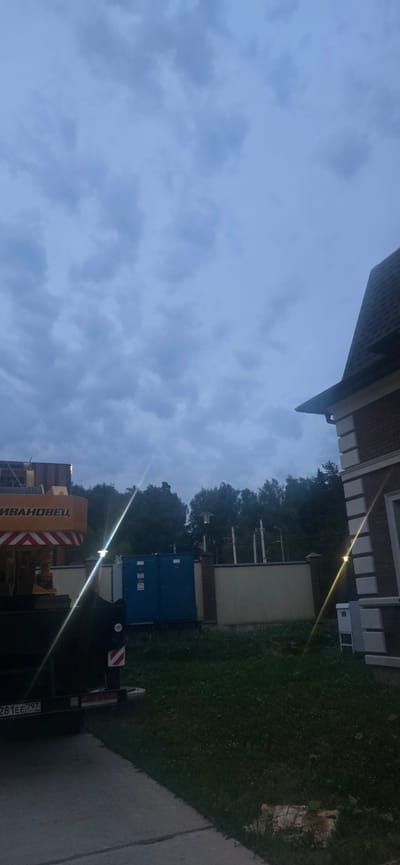 The image depicts an outdoor scene under a cloudy sky, likely in the evening or during overcast weather. In the foreground on the left, a dark-colored vehicle is visible, with a license plate reading "281EE 797". Part of a yellow crane with red and white striped safety markings is also present on the left side of the frame.
In the mid-ground, a blue electrical enclosure or transformer substation is situated behind a light-colored wall. The wall is topped with a decorative brick pattern. Behind the substation and wall, a dense line of trees forms a backdrop.
On the right side of the image, the corner of a brick house with a dark roof is visible. The house features white decorative trim around its windows and along the roofline. A white electrical box is mounted on the exterior wall of the house.
The ground in front of the substation and the house is covered in grass, with some evidence of scattered debris or foliage. A concrete surface, possibly a driveway or pathway, is in the lower left corner of the image. Streaks of light, possibly from artificial illumination or lens flare, are visible across the scene, particularly a bright diagonal streak extending from the lower left to the upper right. The overall impression is an industrial or utility area adjacent to a residential property. The location provided, Русская Деревня-2 Кп, Russia, suggests this is likely a rural or suburban setting with organized housing and potentially shared infrastructure.