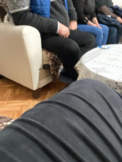 The image displays an indoor scene in the city of Branichevo, Bulgaria, captured from a low, first-person seated perspective. In the immediate foreground, the viewer's legs, clothed in dark, longitudinally ribbed fabric, are prominent. To the left, a segment of a light-colored (off-white) upholstered sofa with dark brown wooden legs is visible. Seated on this sofa is an individual wearing a dark long-sleeved top with patterned light-colored cuffs, a blue puffer vest worn over it, and black pants. Their right hand, adorned with a ring, rests on their lap. A brown and cream patterned blanket or throw is partially visible beneath their thigh. To the right of this individual, at least three other people are seated in a row. One person wears a dark top and blue jeans, while another further right is dressed in dark attire. A white table, draped with a white, patterned lace or embroidered tablecloth, occupies the mid-ground to the right, and a small, light blue object rests upon it. The floor beneath the furniture is light brown parquet or wood laminate, arranged in a patterned layout.