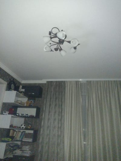 The image depicts an indoor scene, likely a living room or dining area, in Merenii Noi, Moldova. The primary subjects are a white ceiling with a decorative chandelier and a wall with shelves. The chandelier is a dark metal fixture with six white, bell-shaped lamps. Below the ceiling, a wall features a dark-colored shelf unit containing various objects. These include framed pictures or artwork, a clock, several small toy cars, a soccer ball, and books. The background is dominated by a floor-to-ceiling window covered by light gray, vertically pleated curtains. The lighting suggests it is daytime, but the curtains are drawn, indicating either privacy or an attempt to control natural light. The overall impression is of a well-decorated, lived-in domestic space. There is no visible text in the image.