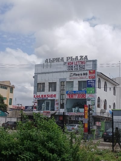 The image shows a white building with a large sign that reads "ALPHA PLAZA NOW LETTING 0806 412 4226 0806 284 5290". The building has multiple storefronts and signs, including "LOLESKIDE" "EXTRA POWER TRINITY DOORS" and "FLOORS NG". There are several cars parked in front of the building. The scene is taken from a street level perspective, with a fence in the foreground and lush vegetation. The sky is overcast with wispy clouds. The location is in Road, Nigeria, at the coordinates 6.439 latitude, 3.52 longitude.