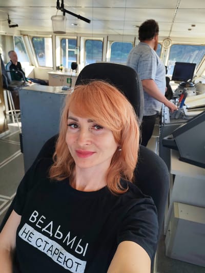 The image depicts the interior of a ship's bridge. In the foreground, a woman with reddish-blonde hair is seated in a chair, looking at the camera and smiling. She is wearing a black t-shirt with white text that reads "ВЕДЬМЫ НЕ СТАРЕЮТ" (Witches do not grow old). Behind her, a man with dark, curly hair and a beard, wearing a grey t-shirt, stands near a control panel. Another person, partially visible and wearing a dark green jacket, is seated to the left. The bridge features large windows offering a view of a body of water and trees on the distant shore, suggesting a riverside or lake setting. The environment appears functional, with various navigational equipment and furniture typical of a ship's bridge. The lighting suggests daytime. The presence of a ship's bridge implies a location associated with water transportation, potentially near or within the city of Pesochnoe, Russia, which is known for its water bodies.