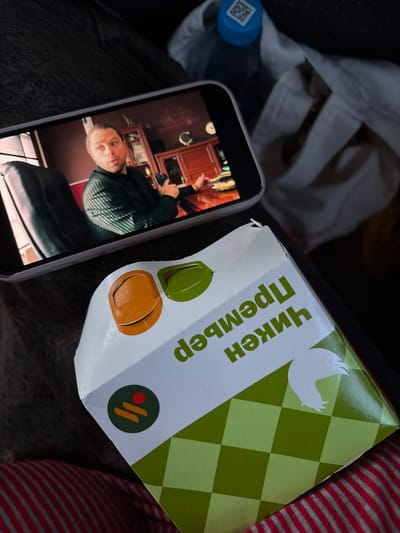 The image shows a person holding a smartphone displaying a video of a man, likely a selfie or live stream, next to a "Chicken Premier" food box written in Cyrillic on what appears to be the interior of a vehicle. There is also a bottle of water and a white cloth bag in the background. There is a red and gray striped seat. The location is likely in Usolye-Sibirskoye, Russia based on the text on the box.